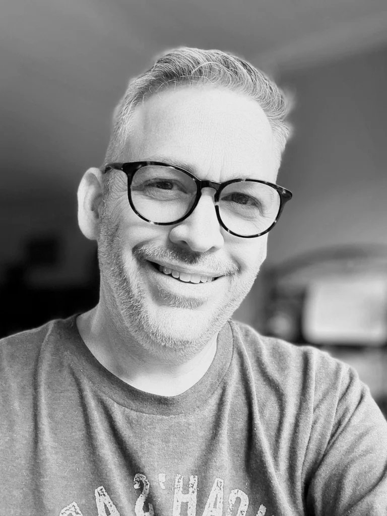 Clay Cartwright smiling in a black-and-white headshot with glasses in a casual workspace.